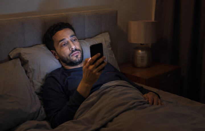 رجل متعب يستخدم هاتفه في السرير ليلاً في صورة تعبر عن الشاشات واضطرابات النوم